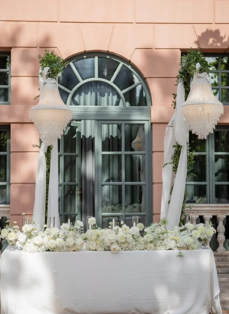 Lámparas de cristal sobre una mesa de flores blancas ante la fachada rosada del hotel