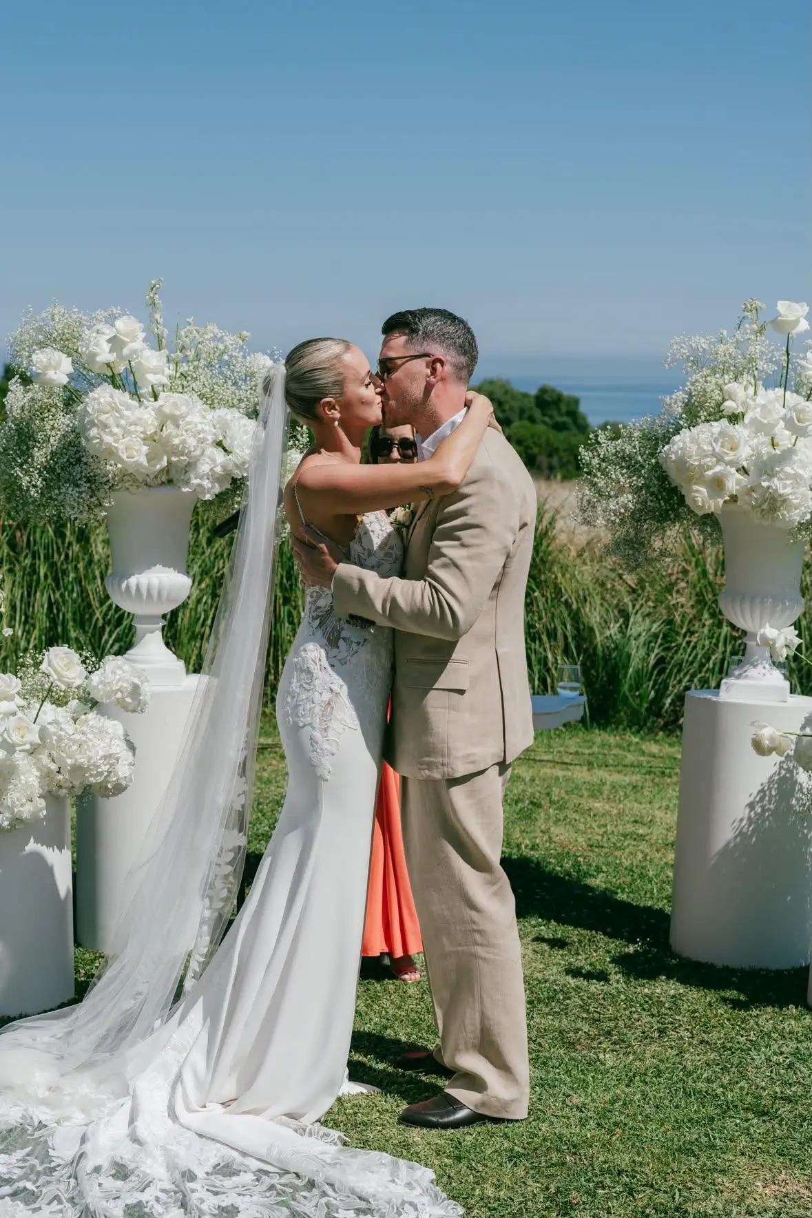 Los novios se besan en la ceremonia, en un jardín con flores blancas y el mar al fondo; la novia lleva un recogido pulido y velo largo