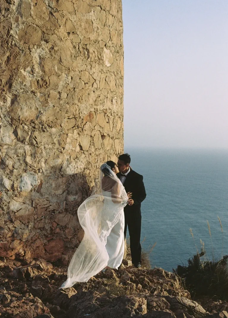 Los novios se besan junto a una torre de piedra, con el velo al viento y el mar detrás