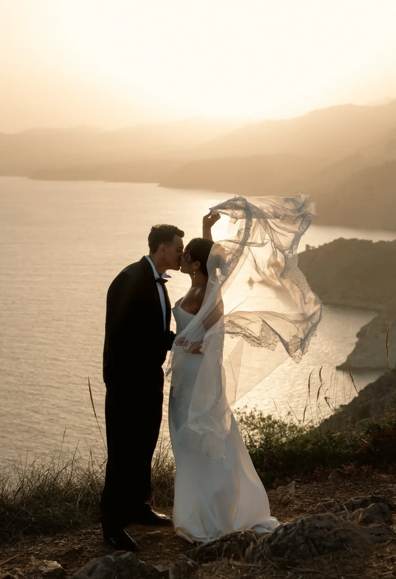 Los novios se besan en lo alto del acantilado, con el velo al viento y la bahía al amanecer