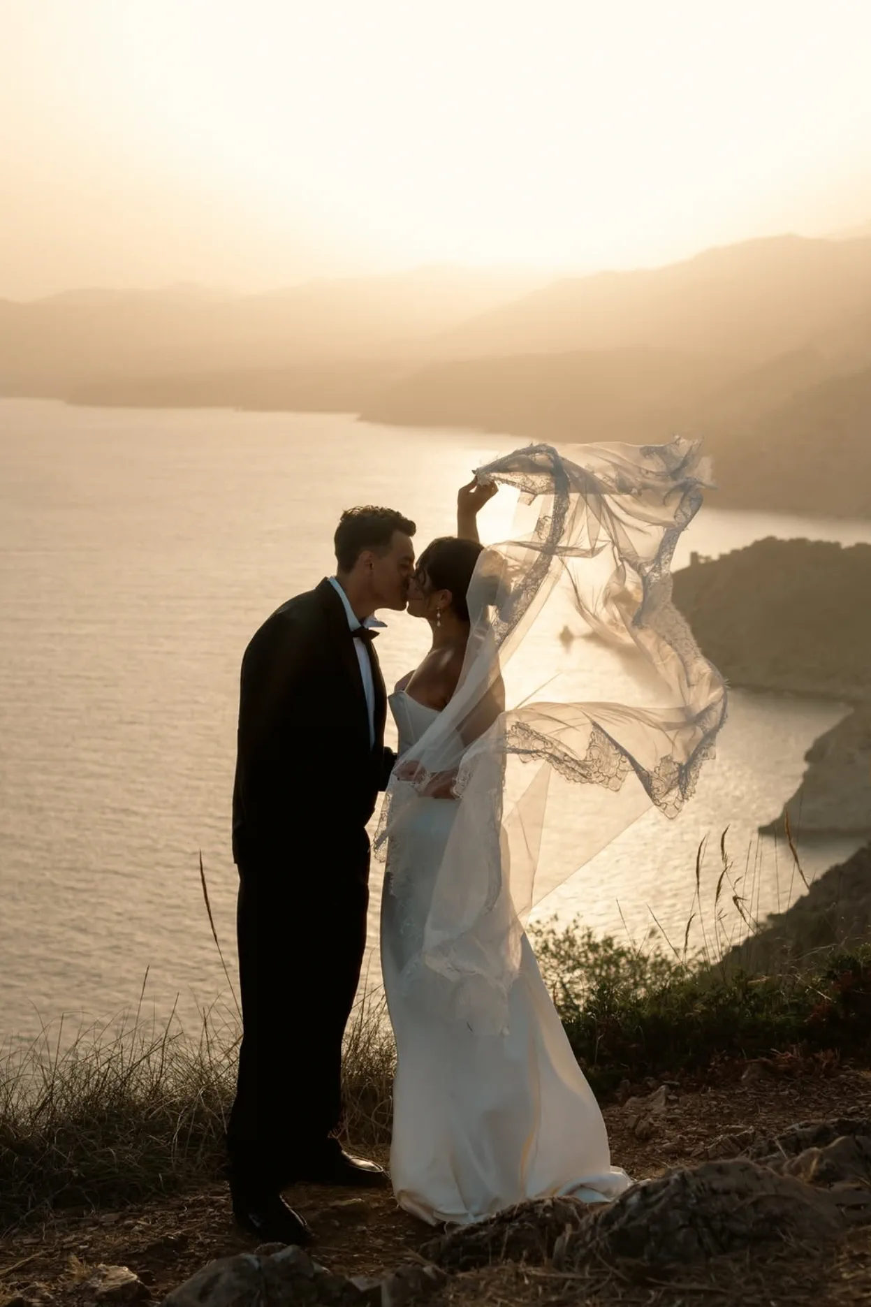 Los novios se besan en lo alto del acantilado, con el velo al viento y la bahía al amanecer