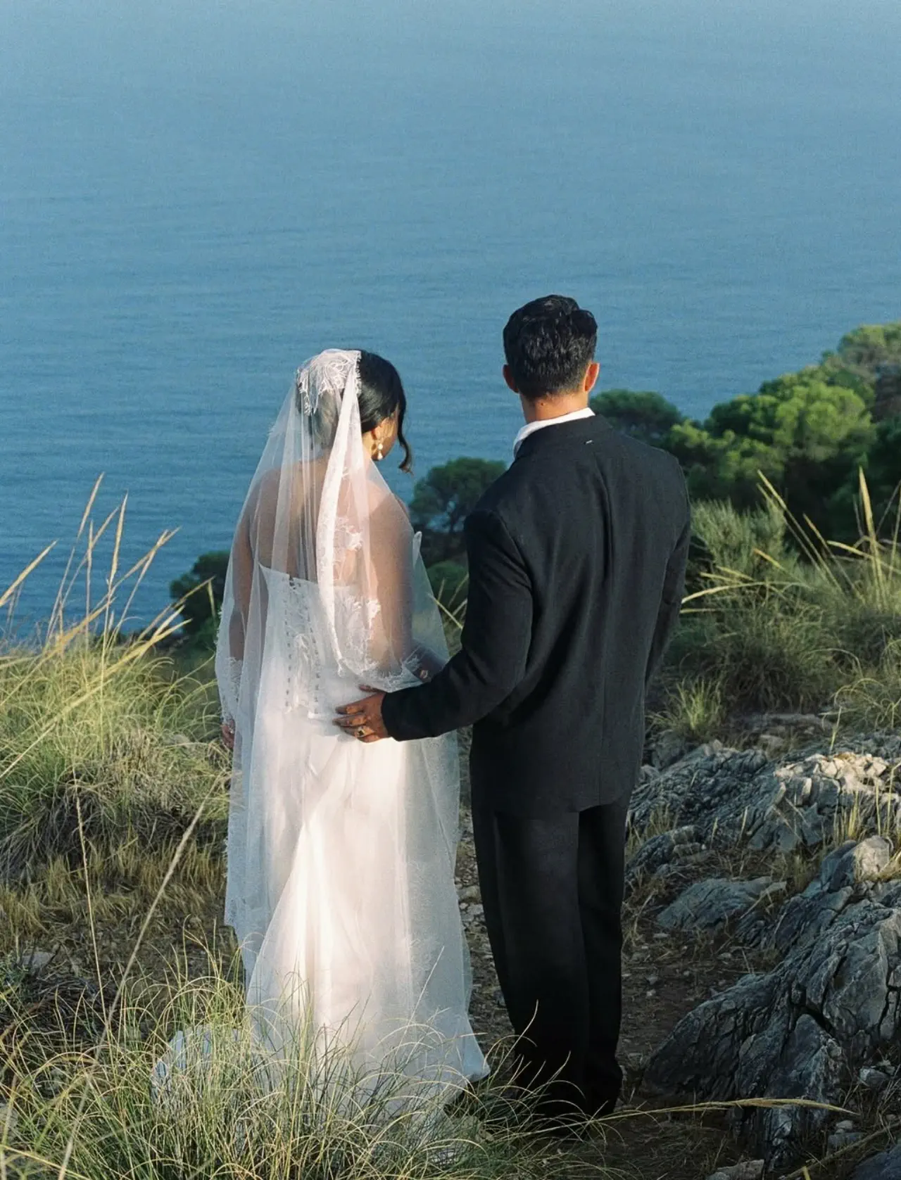 Los novios de espaldas miran el mar; el velo largo cae sobre el recogido de la novia