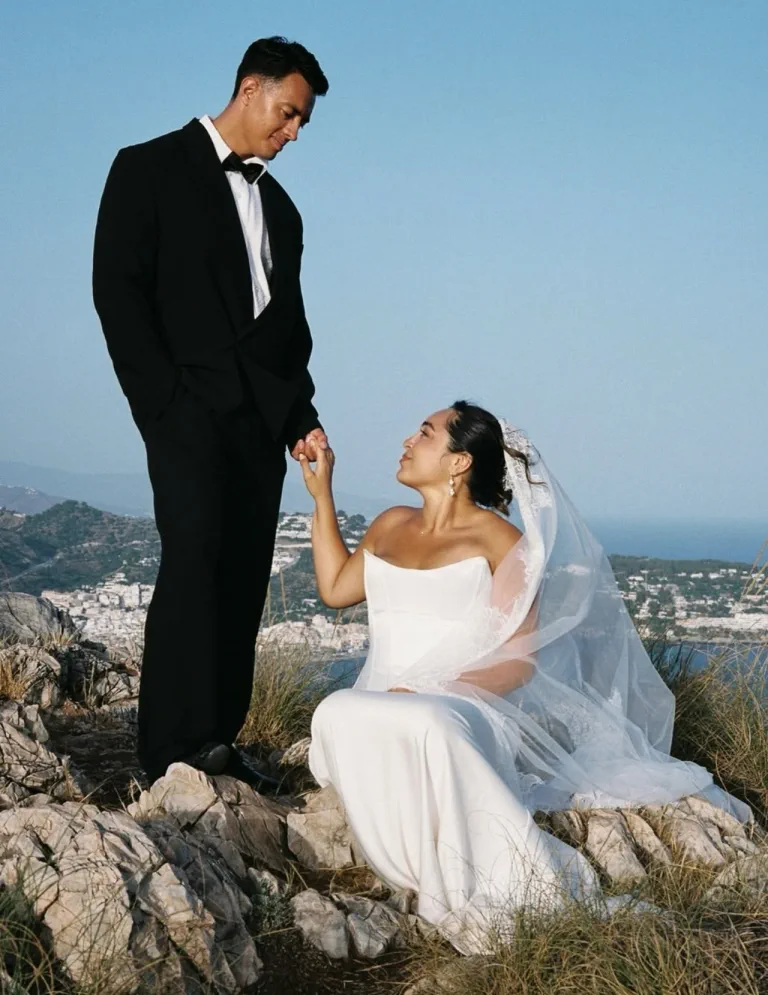 La novia, sentada en las rocas con velo y recogido, da la mano a su marido