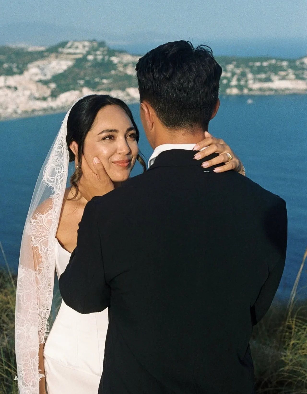 Novia con recogido bajo, velo largo de borde de encaje y maquillaje luminoso, abrazada a su marido ante el mar