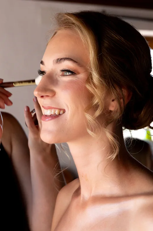 Novia sonriente con maquillaje soft glam en tonos cálidos y recogido bajo con mechones sueltos