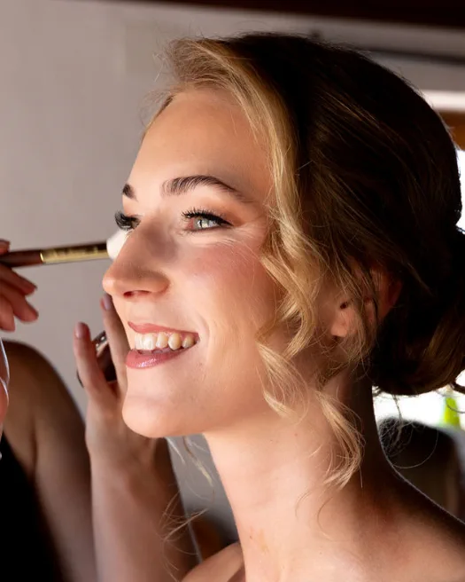 Novia sonriente con maquillaje soft glam en tonos cálidos mientras se aplica el colorete