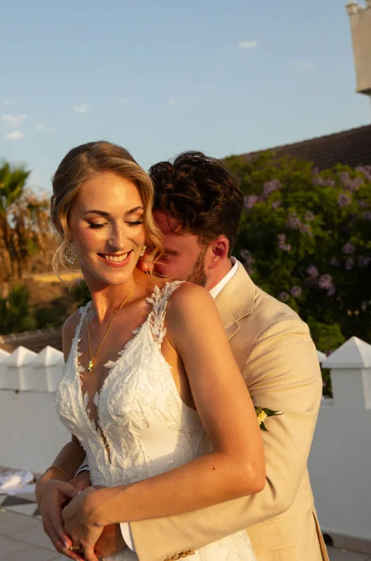 El novio abraza a la novia desde atrás al atardecer en la terraza de Fort Inglés