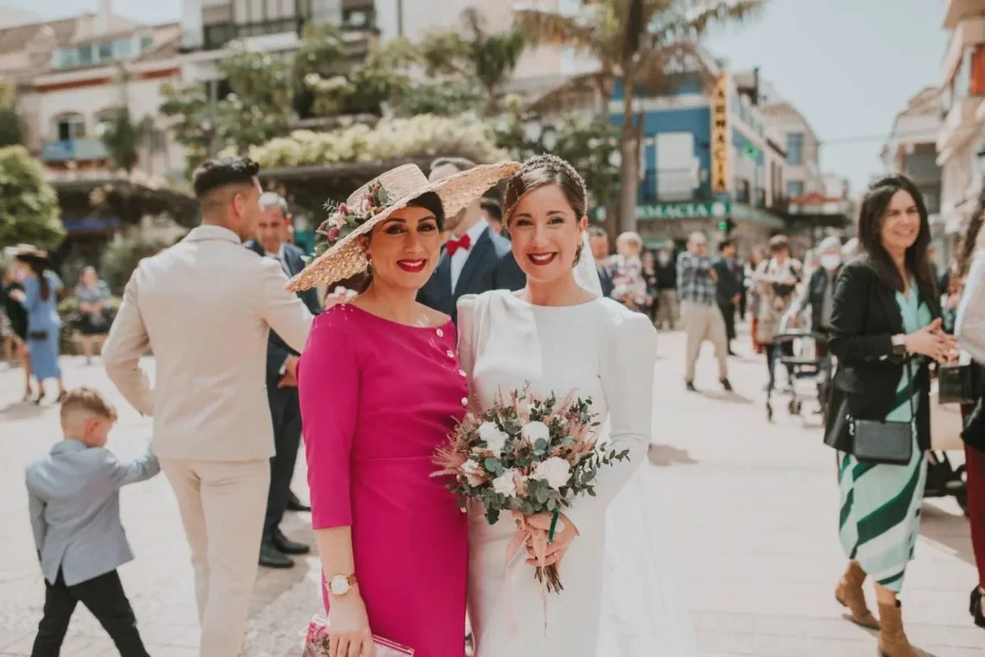 La novia, con su ramo y los labios burdeos, sonríe junto a una invitada en la plaza de la iglesia