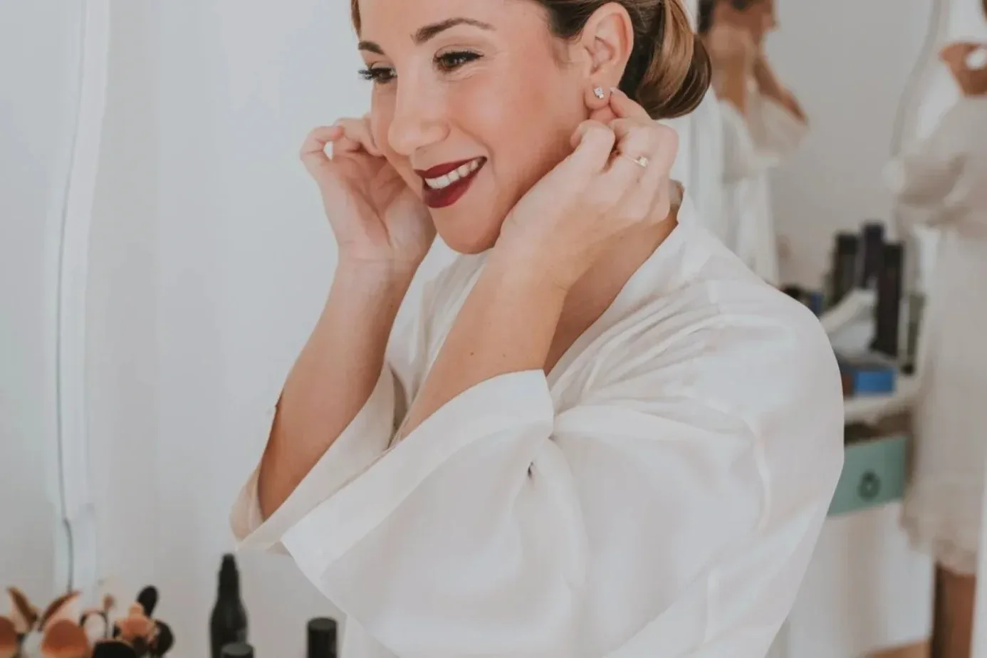 La novia se pone los pendientes, con el recogido pulido y los labios burdeos ya terminados