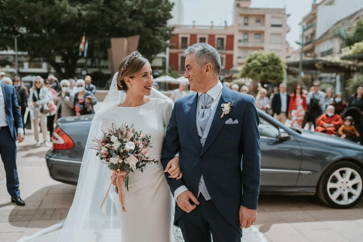 La novia, con velo, recogido pulido y labios burdeos, llega del brazo de un familiar a la iglesia del Rosario