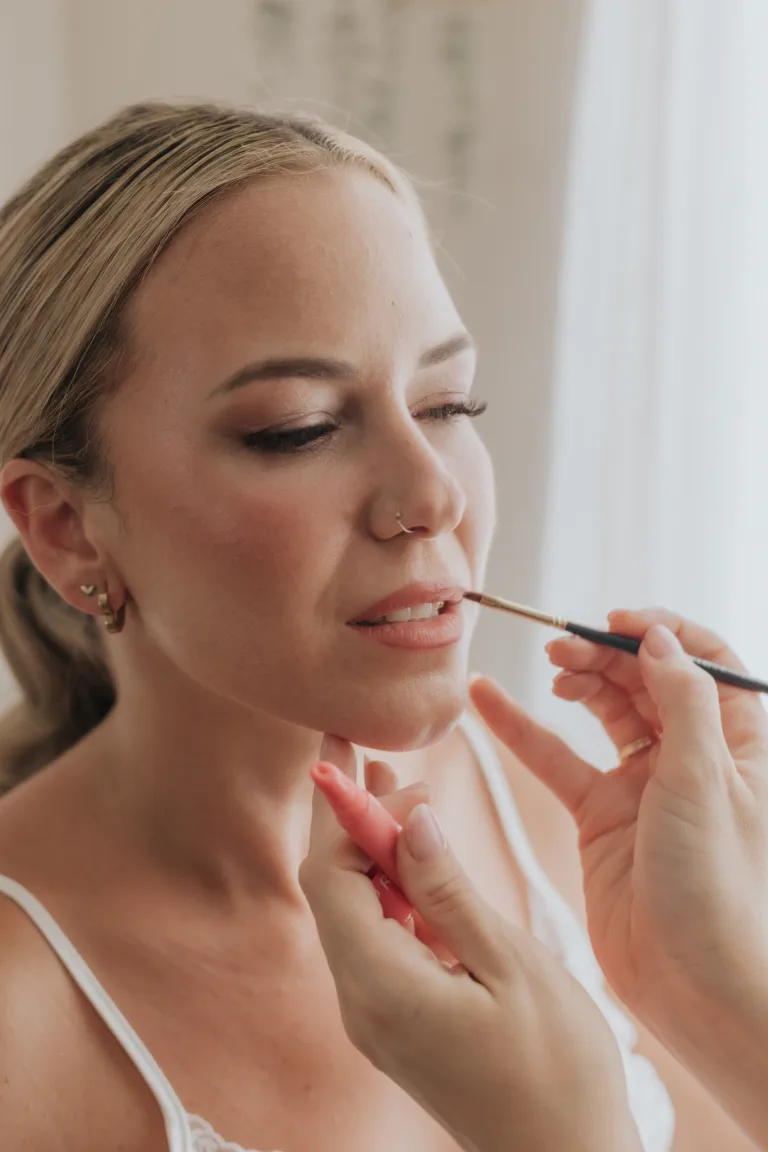 Primer plano de la novia con los ojos cerrados mientras se le perfilan los labios, con un maquillaje soft glam de piel luminosa