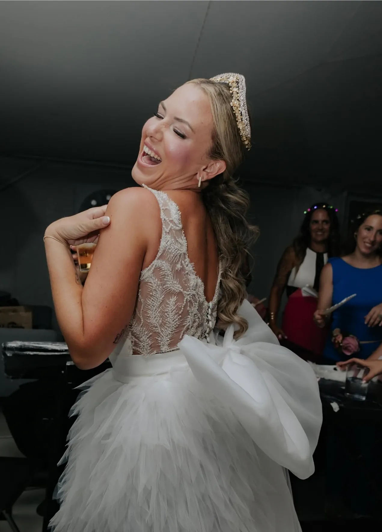 La novia ríe de espaldas en plena fiesta, con tiara dorada y vestido blanco de plumas