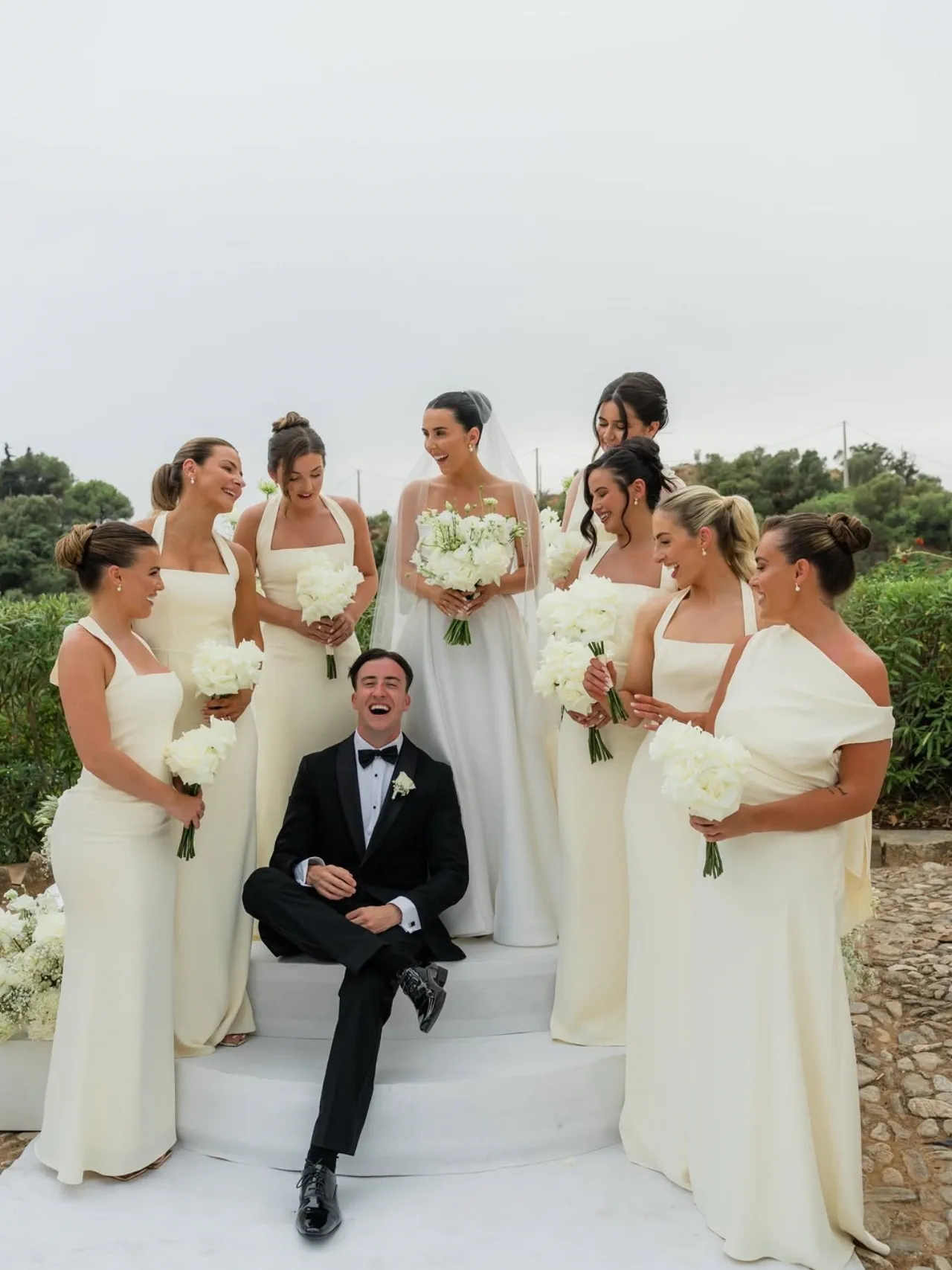 Las damas de honor, con recogidos pulidos, ríen alrededor del novio sentado en una escalera blanca