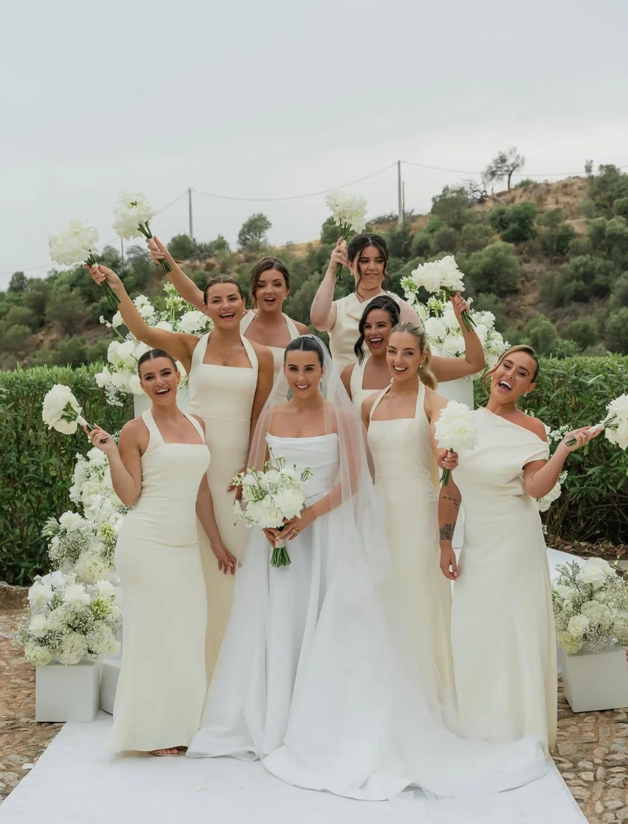 Las damas de honor, con vestidos color crema y peinados pulidos, levantan sus ramos blancos alrededor de la novia