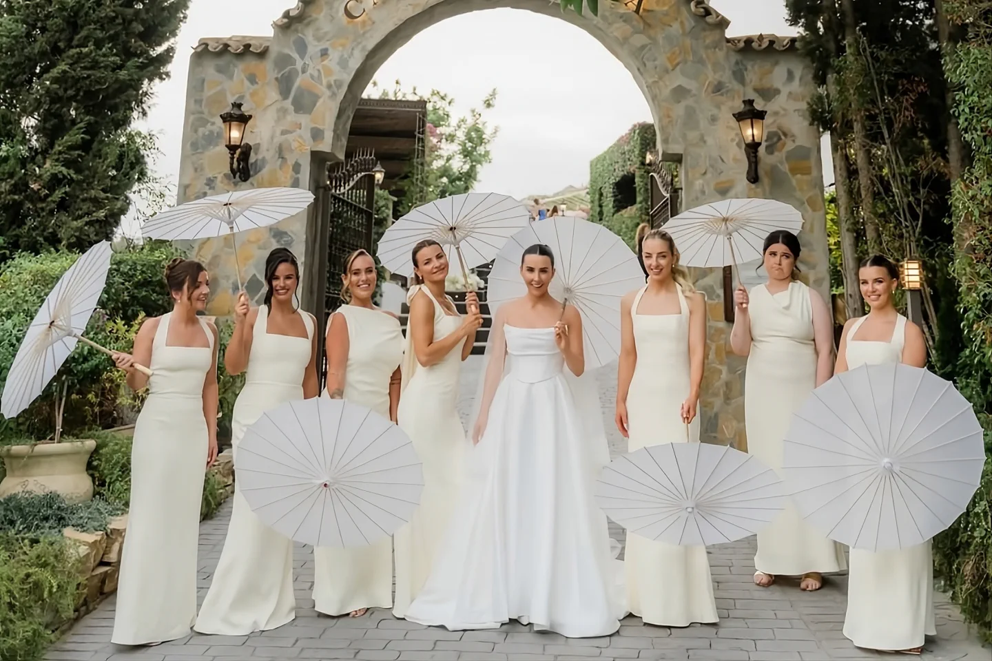 La novia y sus damas de honor con sombrillas blancas bajo el arco de piedra de Casa de la Era